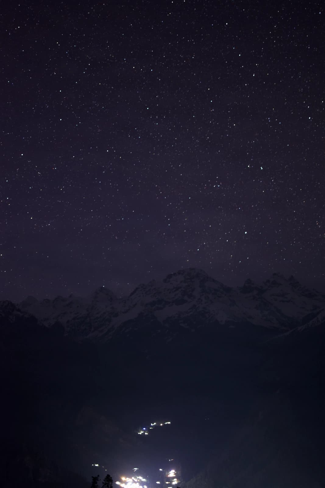 Milky Way arcs above shadowed peaks, a solitary light glimmers below.