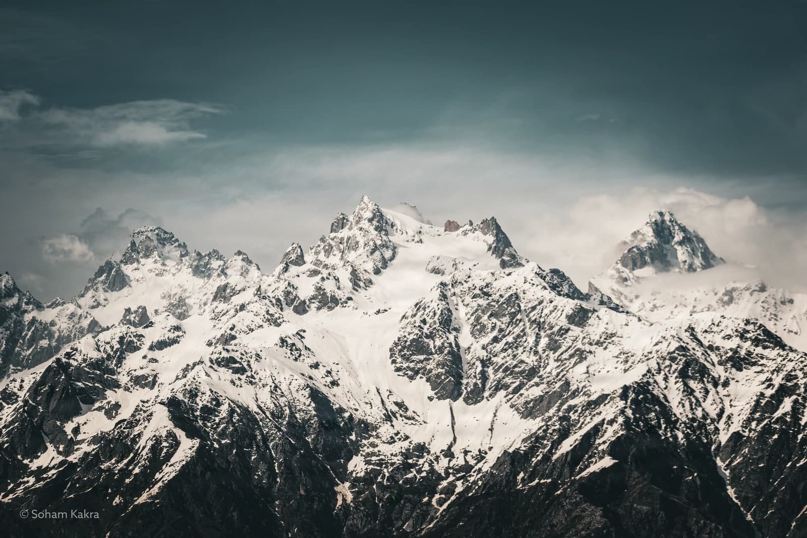 Jagged peaks pierce cloud-laden sky, snow-carved ridges commanding wilderness vastness.