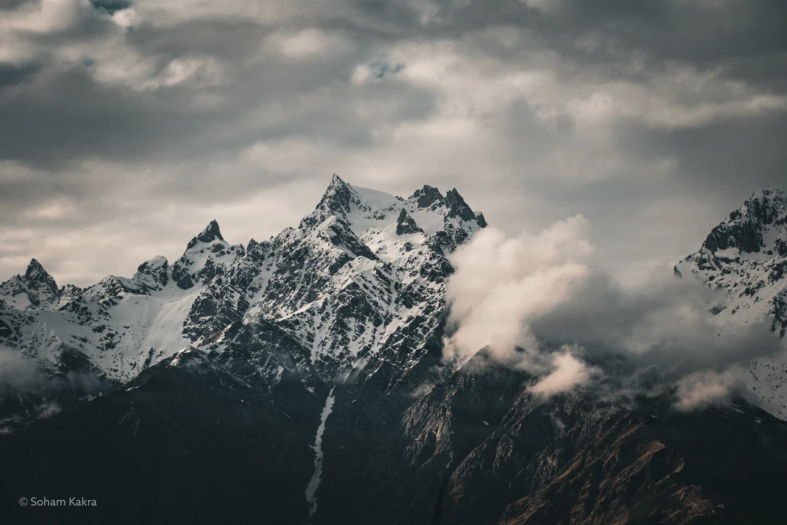 Snow-capped peaks pierce clouds above a forested valley ravine.