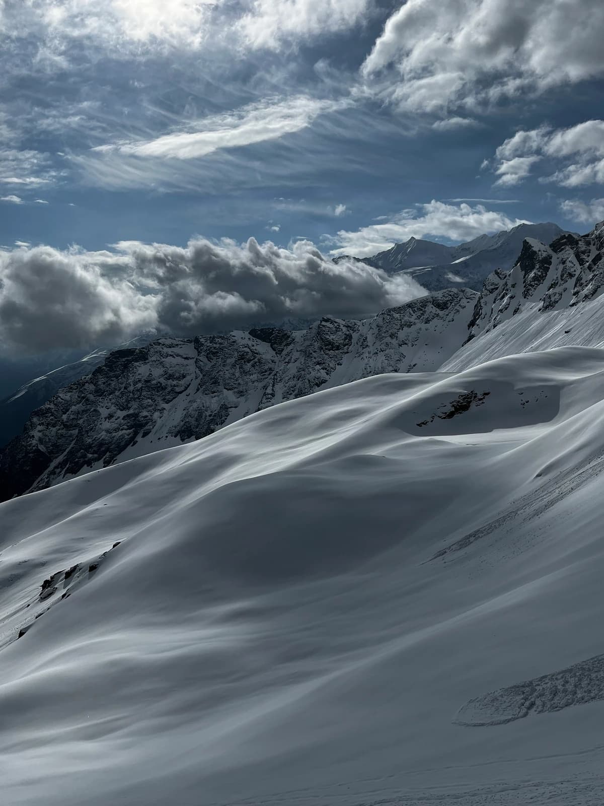 Wind-sculpted snow undulates beneath towering peaks and roiling clouds.